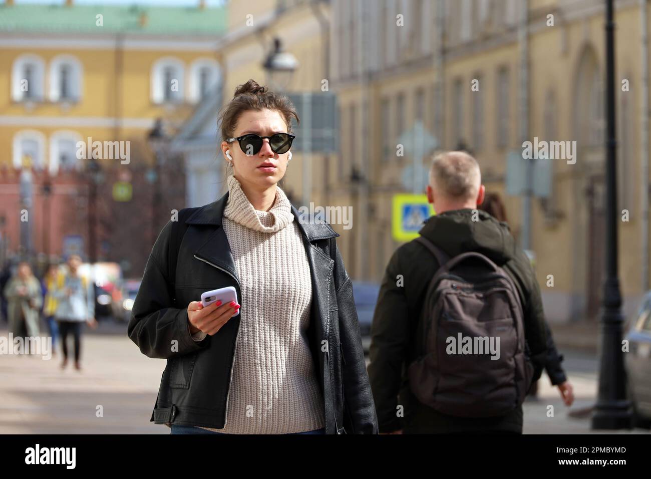 Giovane donna in occhiali da sole e cuffie wireless indossando un cappotto di pelle nera camminando con lo smartphone su una strada sullo sfondo delle persone Foto Stock