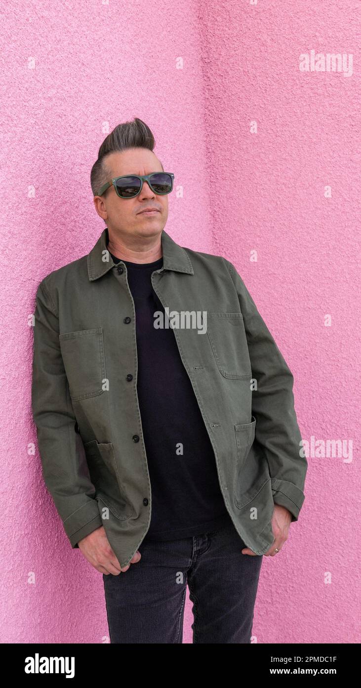 Uomo appoggiato a una parete di stucco rosa in occhiali da sole con giacca verde nera. Foto Stock