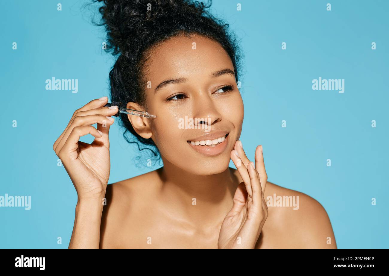 cura della pelle del viso con olio cosmetico e siero per il trattamento. Bella donna africana riccia che applica siero sul viso, isolata su sfondo blu Foto Stock