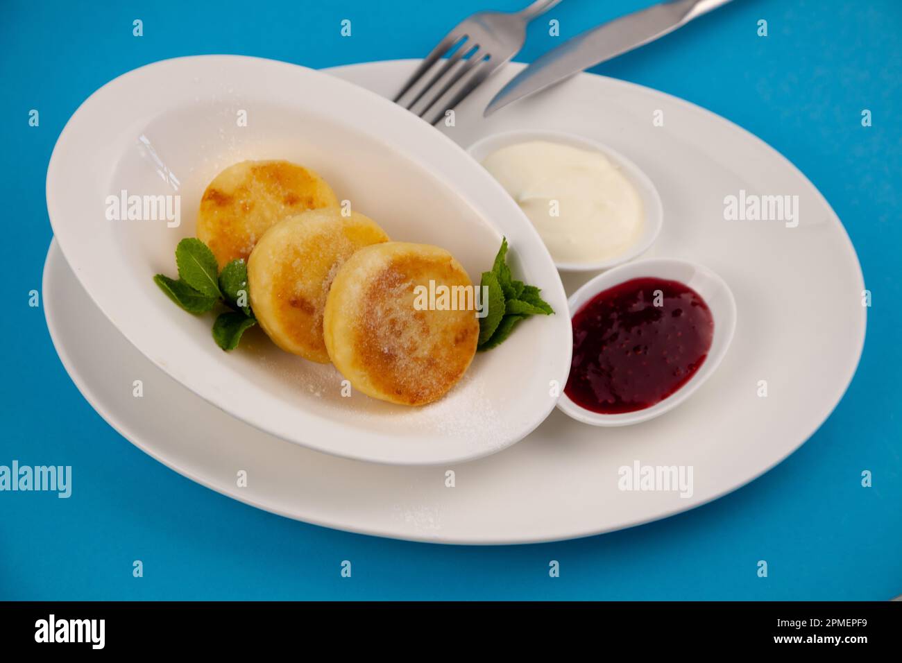 Frittelle di formaggio ricotta rotonda in un piatto bianco su sfondo blu. Cibo prima colazione. Cibo sano. Salsa di panna acida e menta. Primo piano vista dall'alto. Pasto dessert. Frittelle di ricotta al formaggio ricotta. Foto Stock