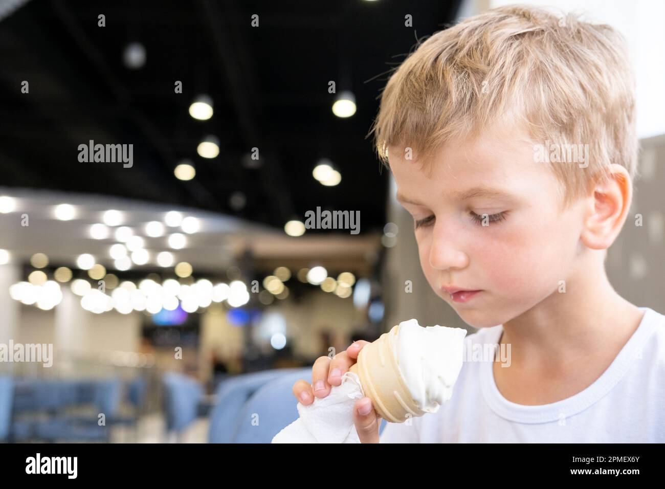 Ritratto di un piccolo ragazzo affamato divertente mangiare gelato freddo delizioso in una tazza di waffle seduti in un caffè, sfondo scuro, bokeh. Un bambino gode di cre di ghiaccio Foto Stock