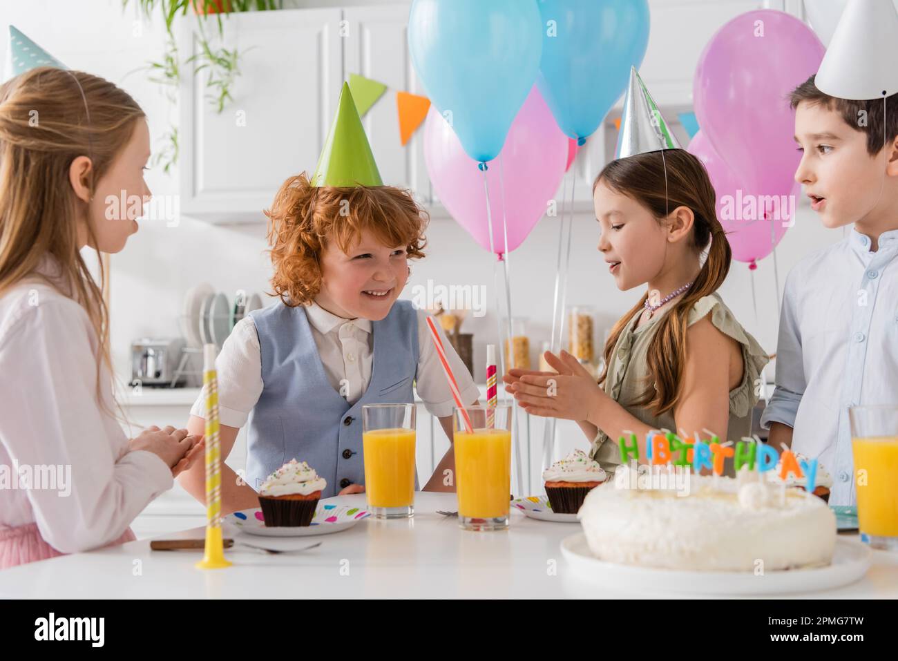 le ragazze della pre in cappellini del partito che batte le mani e cantano la canzone felice di compleanno vicino ai ragazzi e alla torta, immagine di scorta Foto Stock