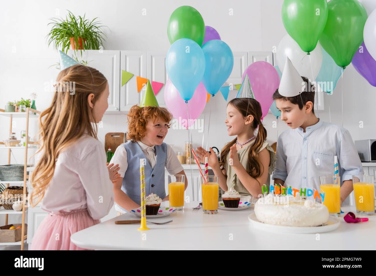ragazze felici in cappellini di festa che batte le mani e cantano canzone di buon compleanno vicino ai ragazzi e torta, immagine di scorta Foto Stock