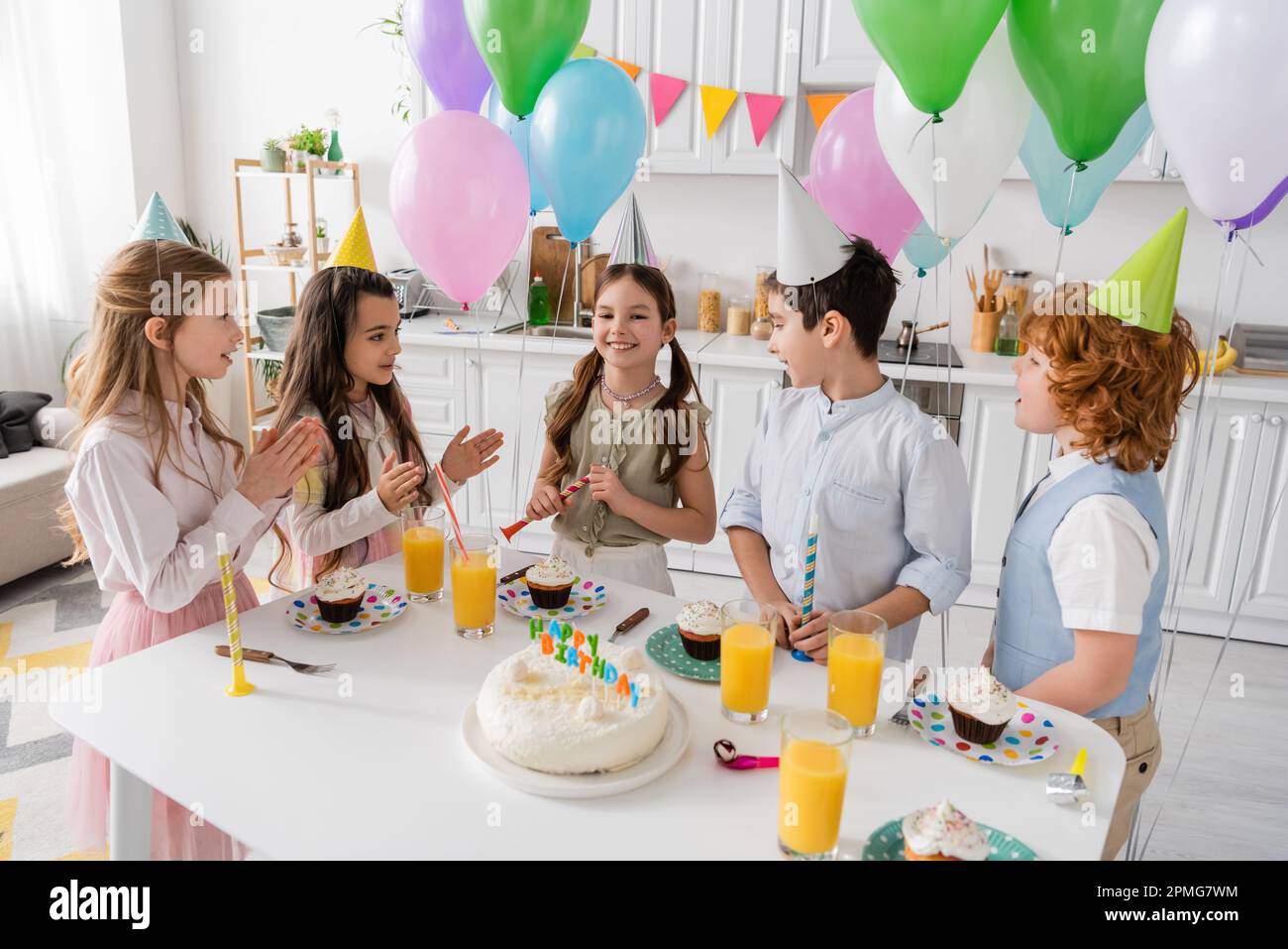 bambini positivi che battono le mani e cantano felice canzone di compleanno accanto alla torta con candele e palloncini, immagine stock Foto Stock