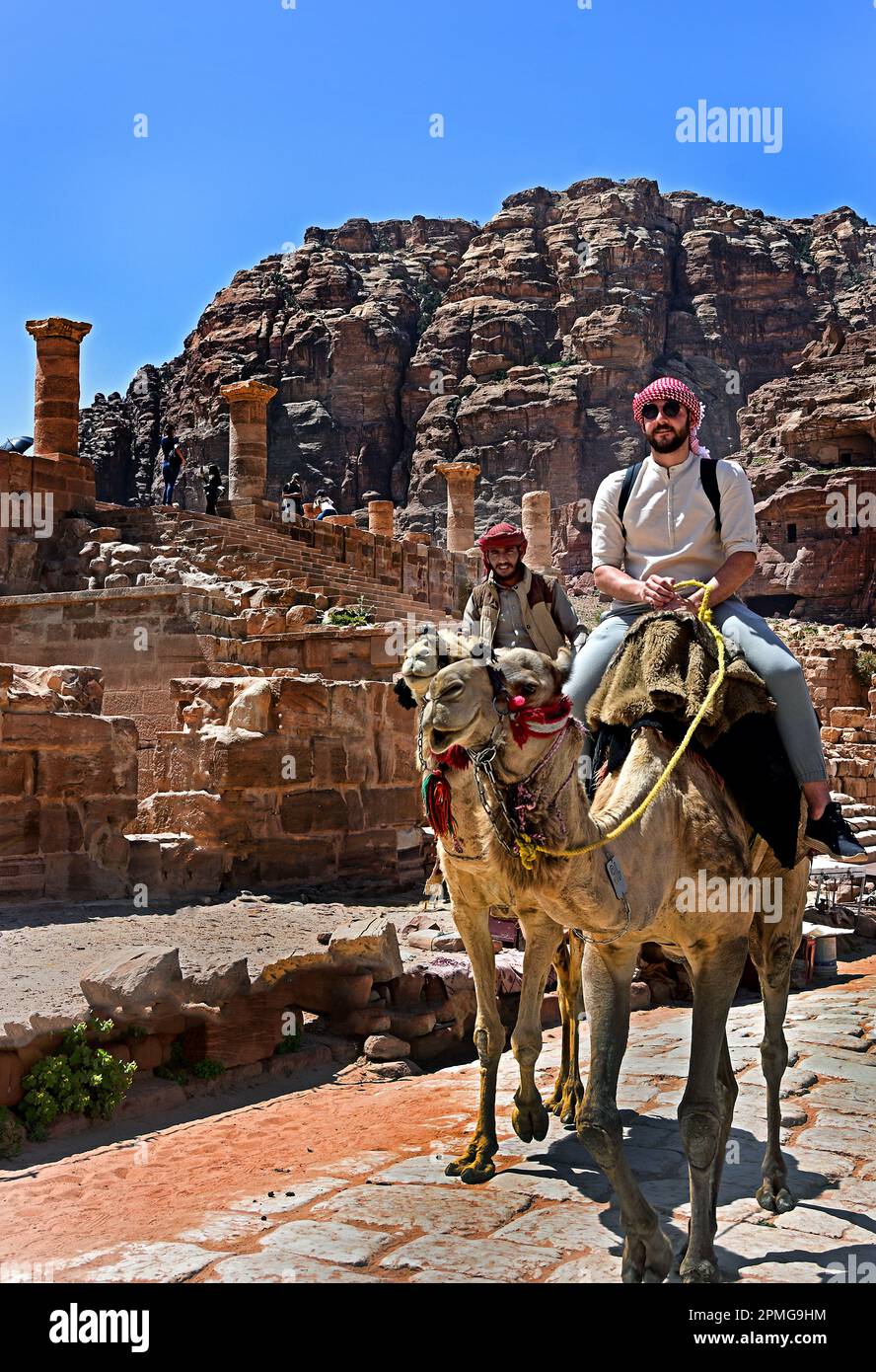 Cammelli e cavalli lungo il sentiero polveroso del deserto sotto la maestosa facciata scavata nella roccia del Tombe del Palazzo nell'antica città nabatea di Petra, Giordania. Foto Stock