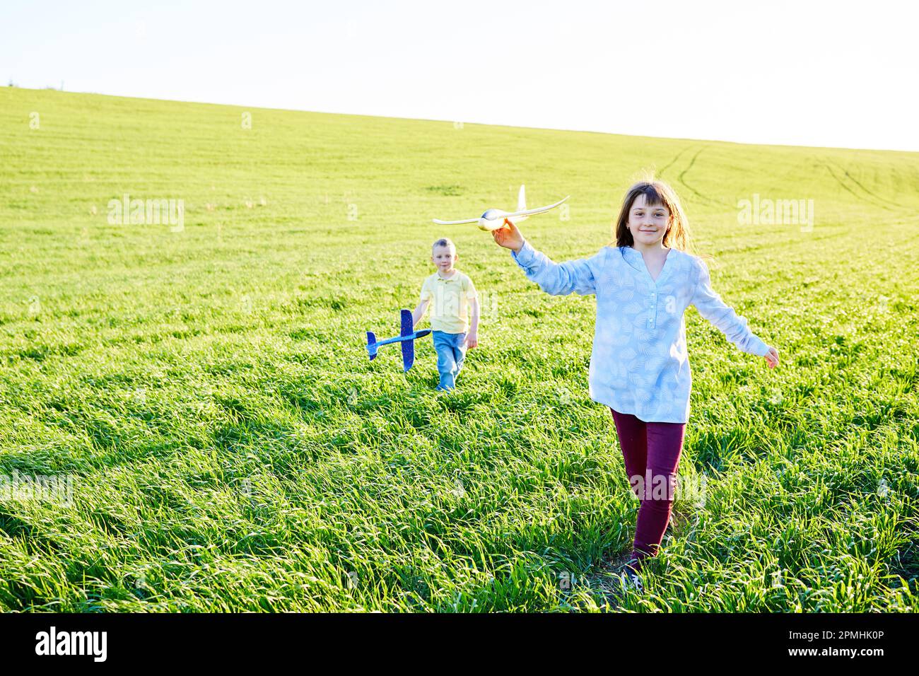 Il ragazzo e la ragazza che corrono che tengono due aeroplani gialli e blu giocattolo nel campo durante la giornata estiva di sole Foto Stock
