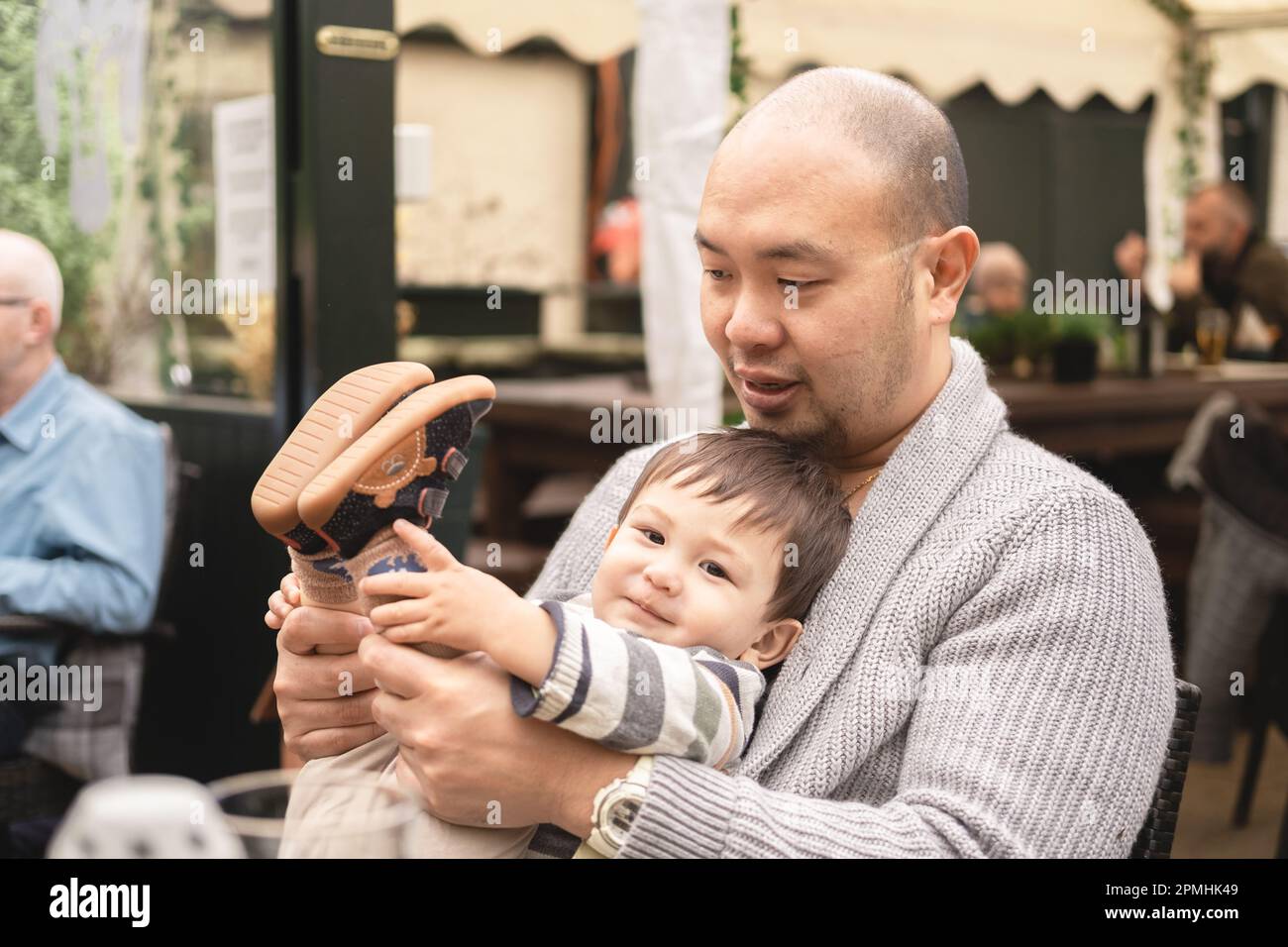 Bambino maschio flessibile che guarda la macchina fotografica seduta sulle gambe del papà, tenendo le gambe, giocando con il padre nella birreria all'aperto al pub in Scozia. Gioioso. Foto Stock