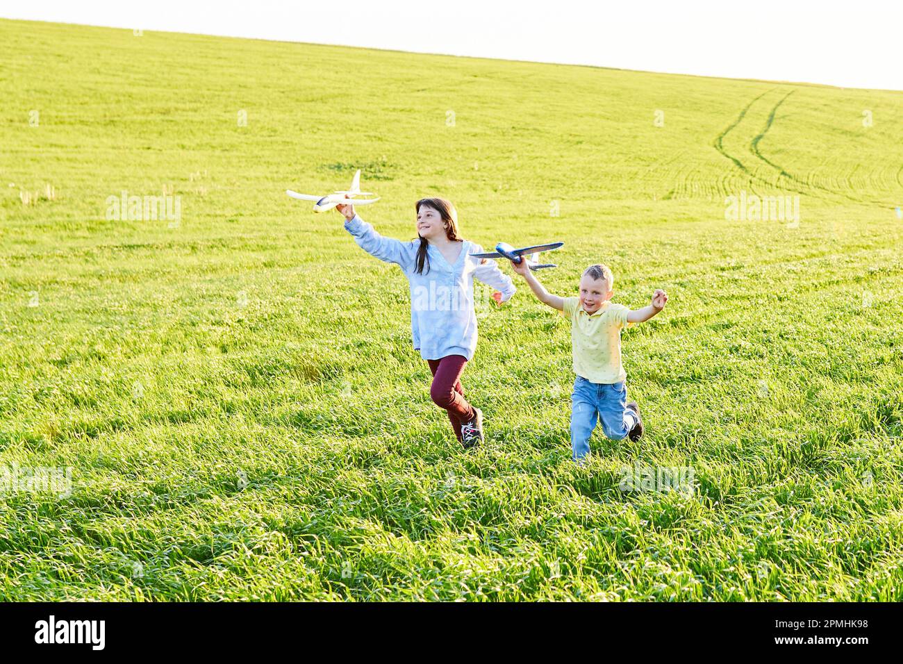 Il ragazzo e la ragazza che corrono che tengono due aeroplani gialli e blu giocattolo nel campo durante la giornata estiva di sole Foto Stock
