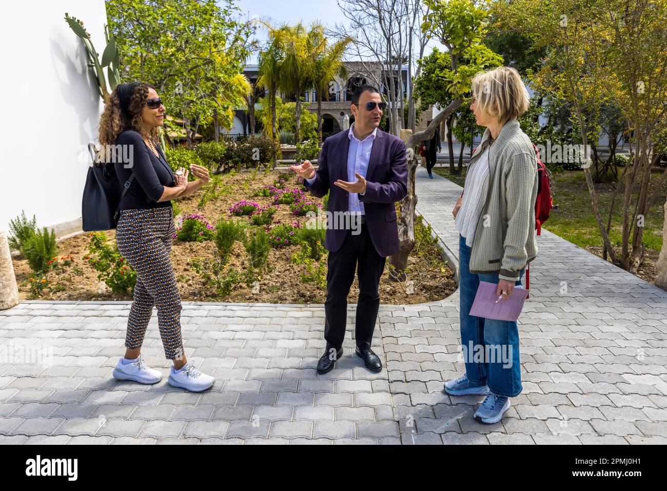 Prof. Dr. Asım Vehbi, rettore dell'Università Arkin di Arti Creative e Design (ARUCAD), parla e gesticola con due visitatori in un giardino nel campus universitario di Kyrenia (turco: Girne), Repubblica Turca di Cipro del Nord (RTNC). A destra, la giornalista Angela Berg ascolta, taccuino in mano. Fondata nel 2017 dal gruppo Arkın, l'università è un'istituzione tematica dedicata esclusivamente all'arte, al design e alla comunicazione. Foto Stock