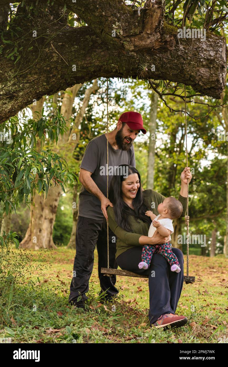 Famiglia felice, padre e madre sorridendo e guardando la bambina, all'aperto. Madre seduta su un'oscillazione con il suo bambino ed il marito in piedi vicino a. Foto Stock