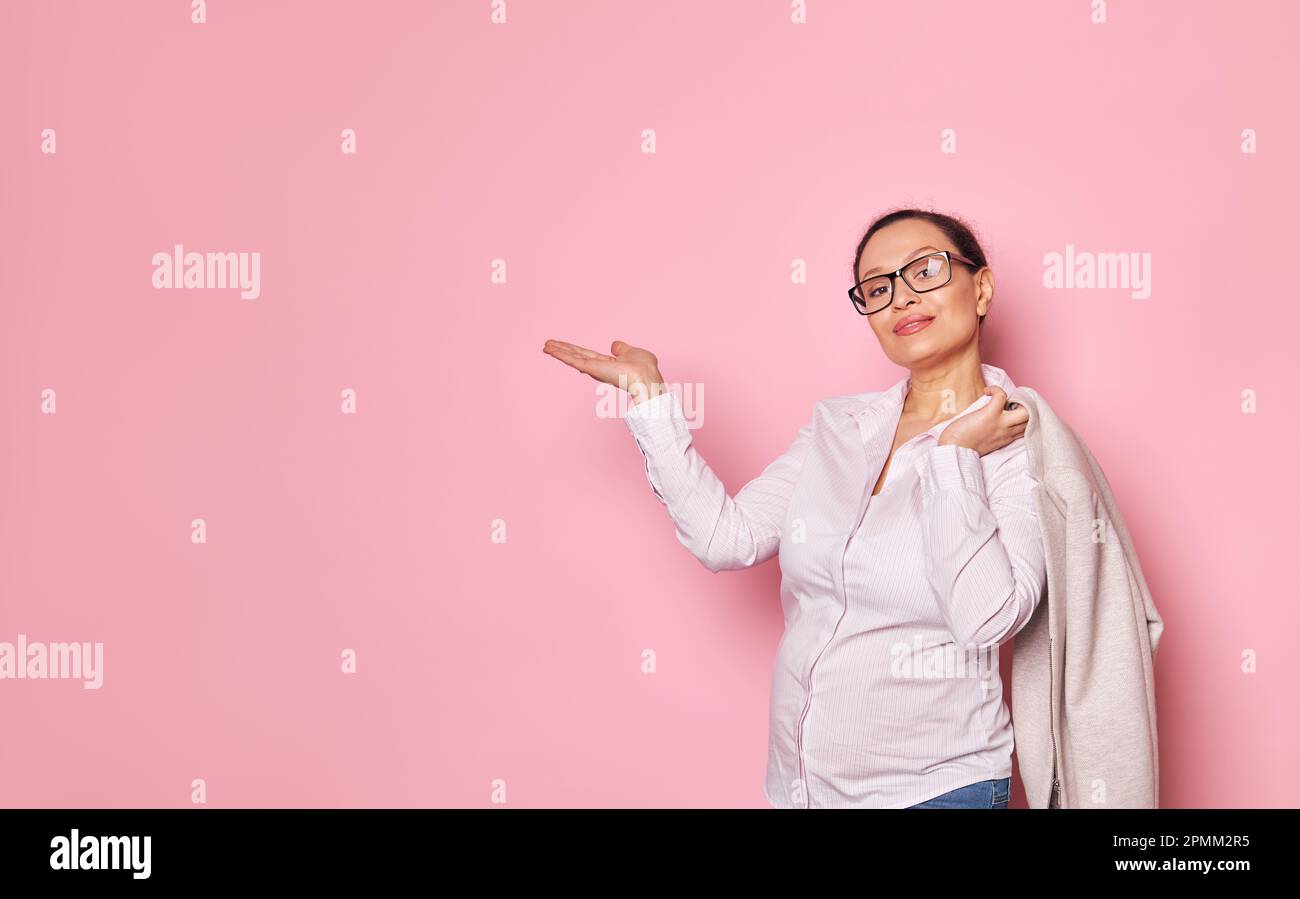 Donna incinta sicura in occhiali alla moda, tenendo spazio copia immaginaria sulla sua mano palmo in su, su sfondo rosa isolato Foto Stock