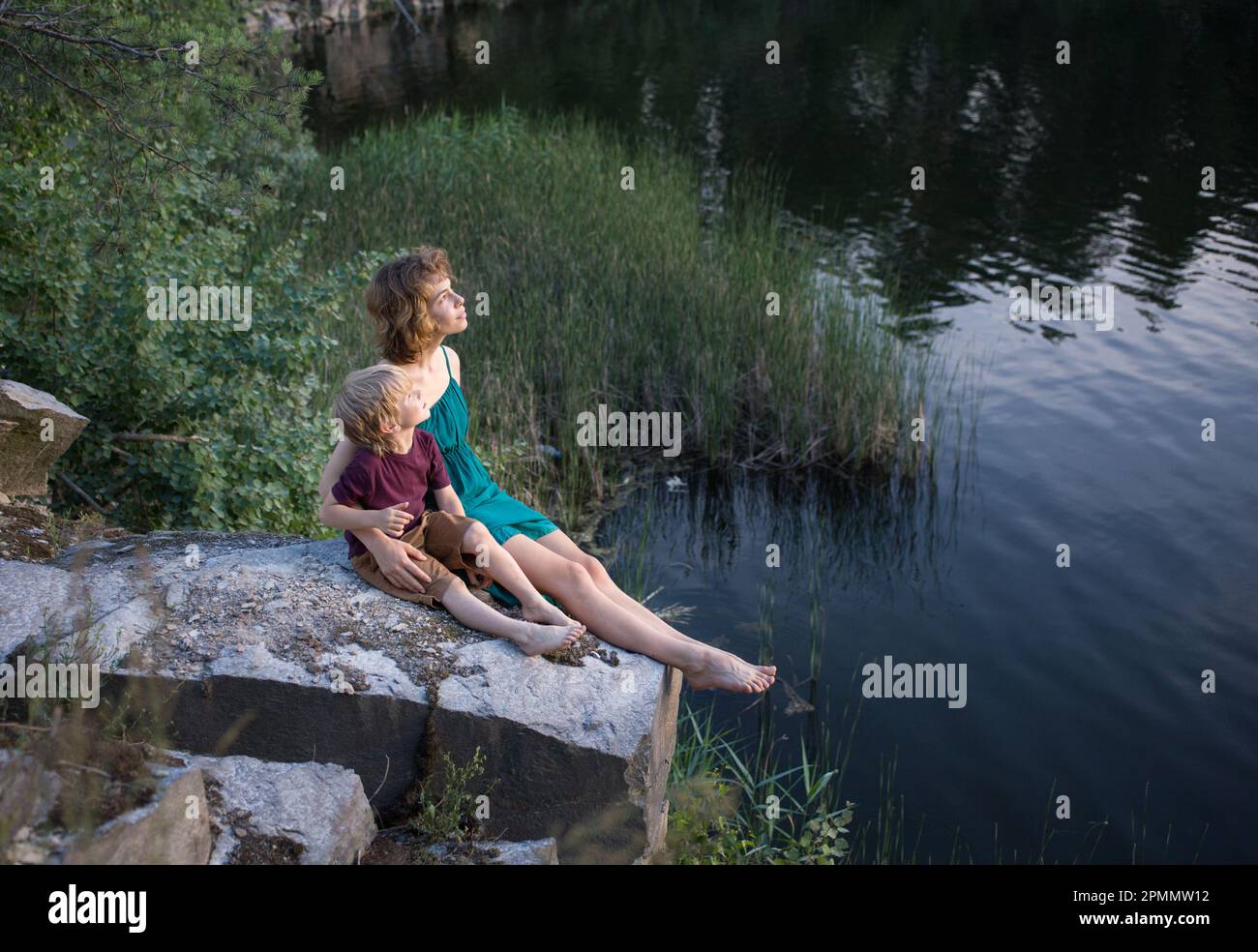 ragazza adolescente di 16 anni e un ragazzo di 5 anni (fratello e sorella) sono felicemente seduti insieme su una roccia vicino al lago e fantasiose con int Foto Stock