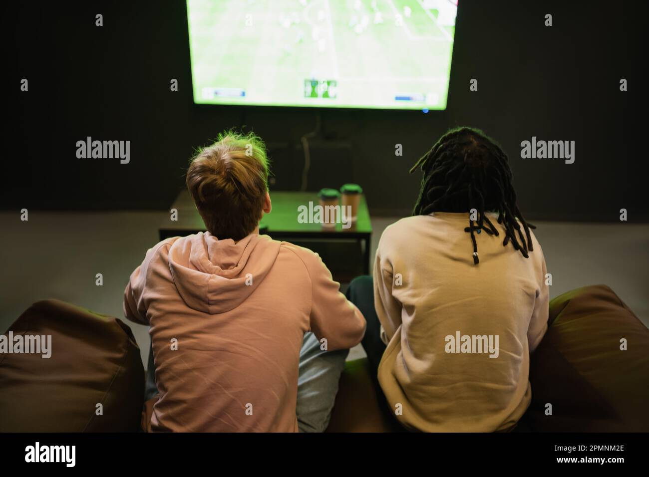 Vista posteriore degli amici interracial che guardano la partita sportiva vicino caffè sfocato per andare in cyber club, immagine stock Foto Stock