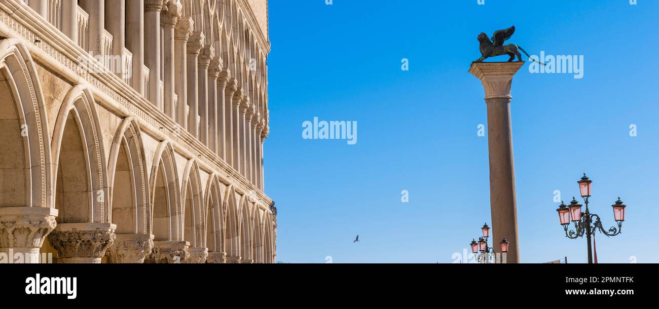 La statua del leone alato in cima alla colonna di San Marco in Piazza San Marco con gli archi bianchi del Palazzo Ducale contro un cielo blu Foto Stock