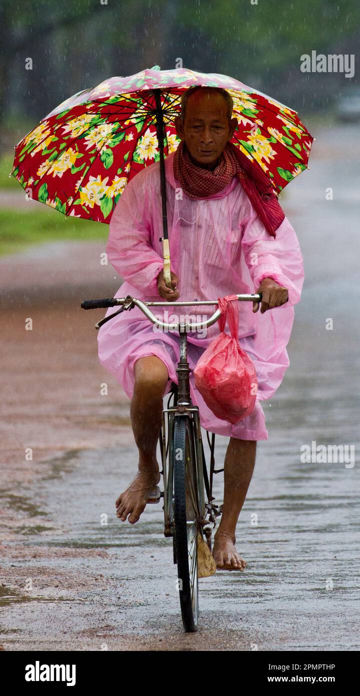 Uomo cambogiano con un ombrello in sella a una bicicletta sotto la pioggia; Siem Reap, Cambogia Foto Stock