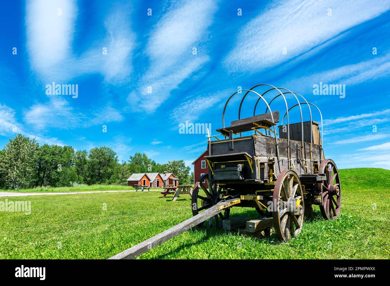 Vecchio carro in legno con spettacolari nuvole e cielo blu con edifici dipinti di rosso sullo sfondo, a sud di Longview, Alberta; Alberta, Canada Foto Stock