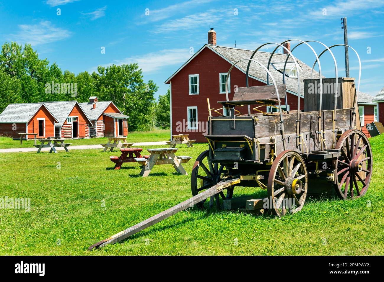 Vecchio carro in legno con edifici dipinti di rosso, tavoli da picnic, cielo blu e nuvole sullo sfondo, a sud di Longview, Alberta; Alberta, Canada Foto Stock