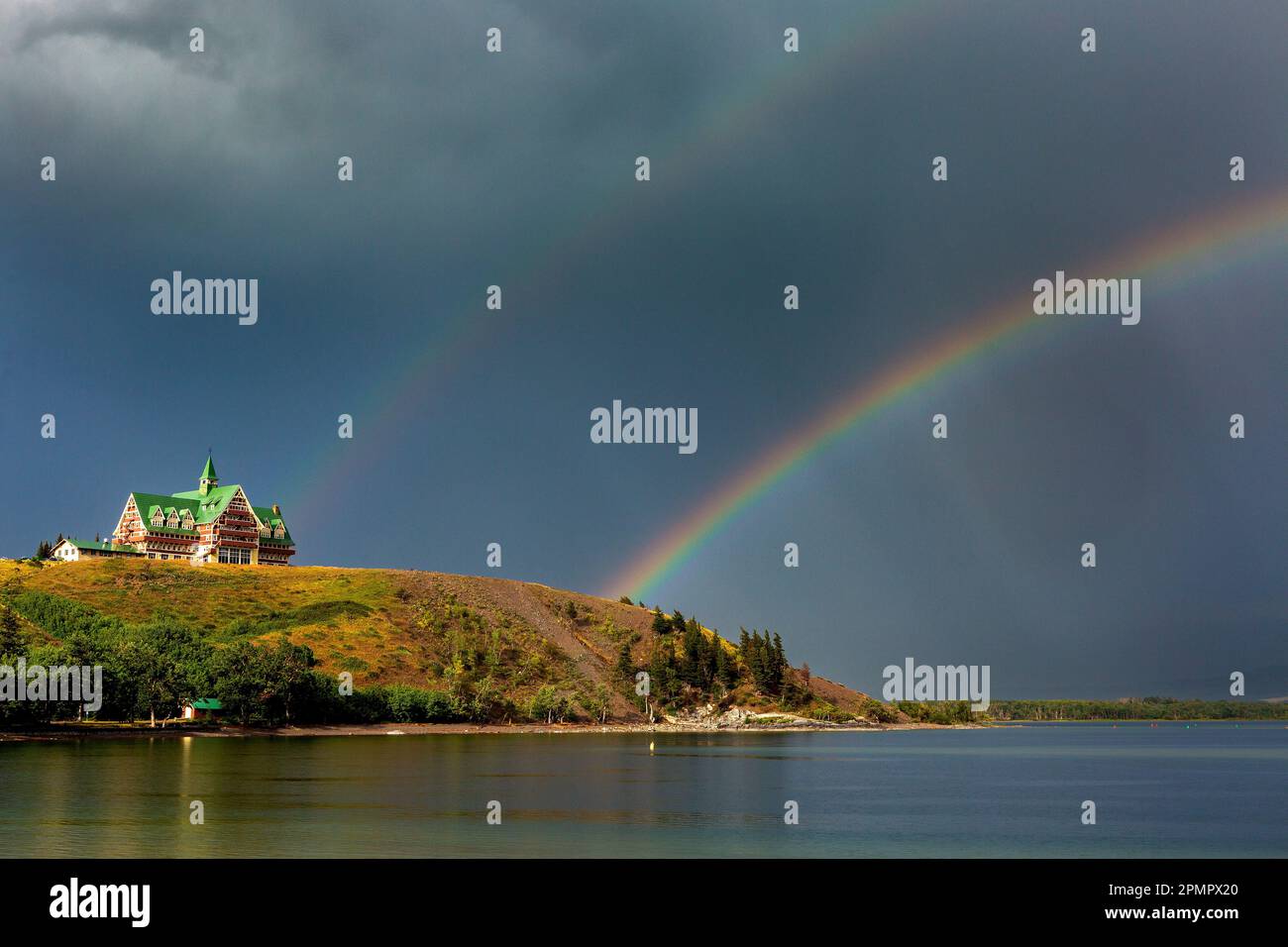 Doppio arcobaleno in un cielo scuro su un lago con uno storico edificio in legno sul fianco della collina che si affaccia sul lago, che si illumina di luce calda al tramonto Foto Stock