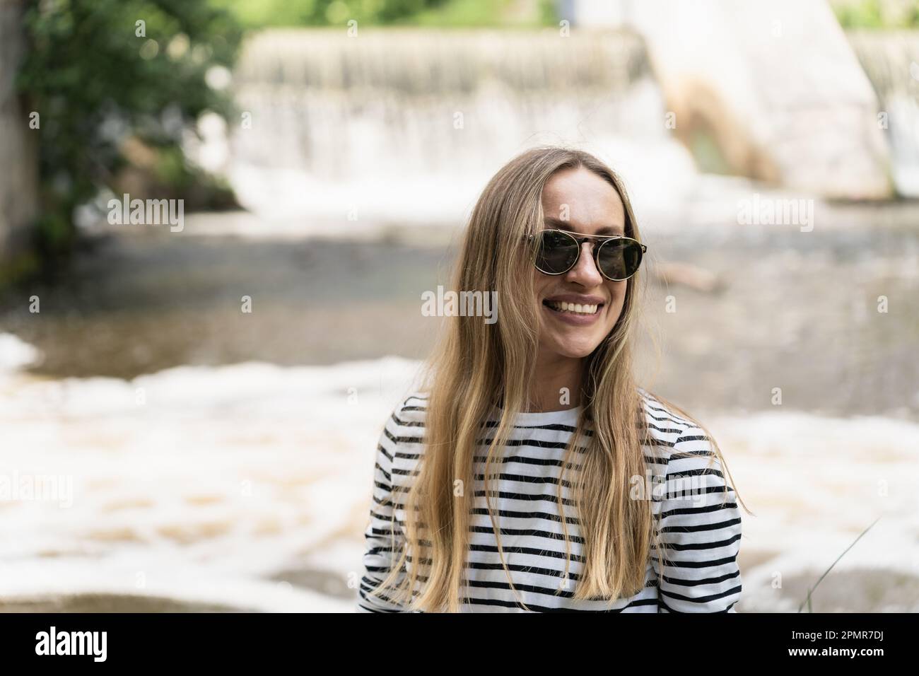 Donna sorridente dai capelli lunghi in occhiali da sole vicino al fiume di fronte alla diga. Foto Stock
