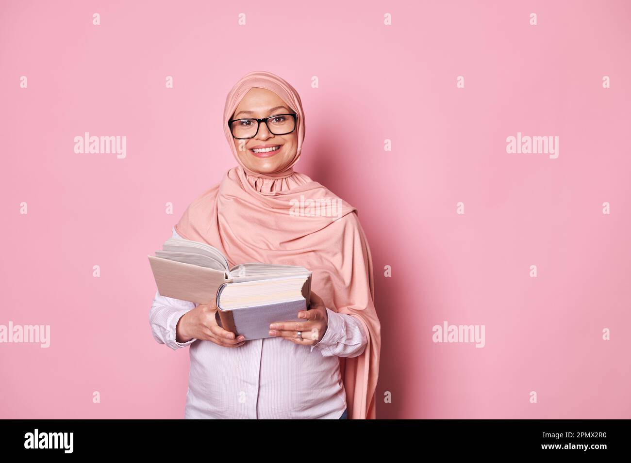 Sicura bella donna musulmana incinta in hijab rosa e gli occhiali alla moda, leggendo un libro, sorridendo guardando la macchina fotografica Foto Stock