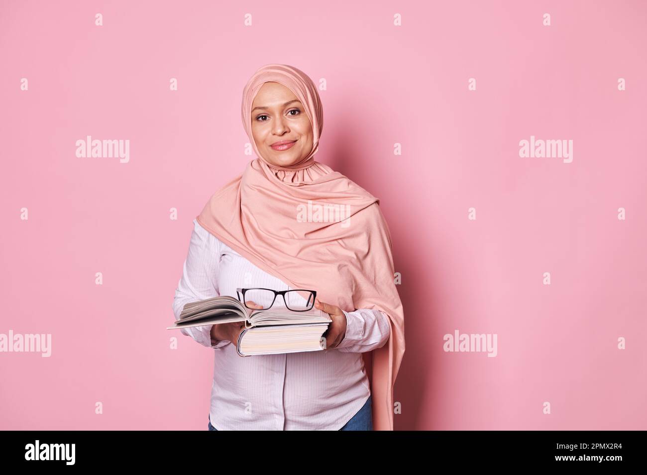 Donna musulmana fiduciosa educatore in hijab rosa, un insegnante di scuola che tiene occhiali e libro di studio, sfondo rosa Foto Stock