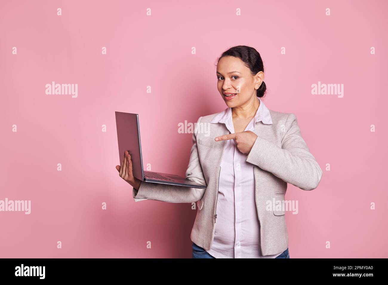 Charming African American business donna manager punti a computer portatile, sorride guardando la fotocamera, isolato su rosa Foto Stock