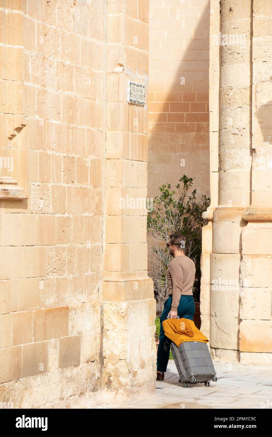 Mdina, Malta - 13 novembre 2022: Uomo che cammina per la strada pedonale calcarea mentre tira la valigia, da dietro Foto Stock