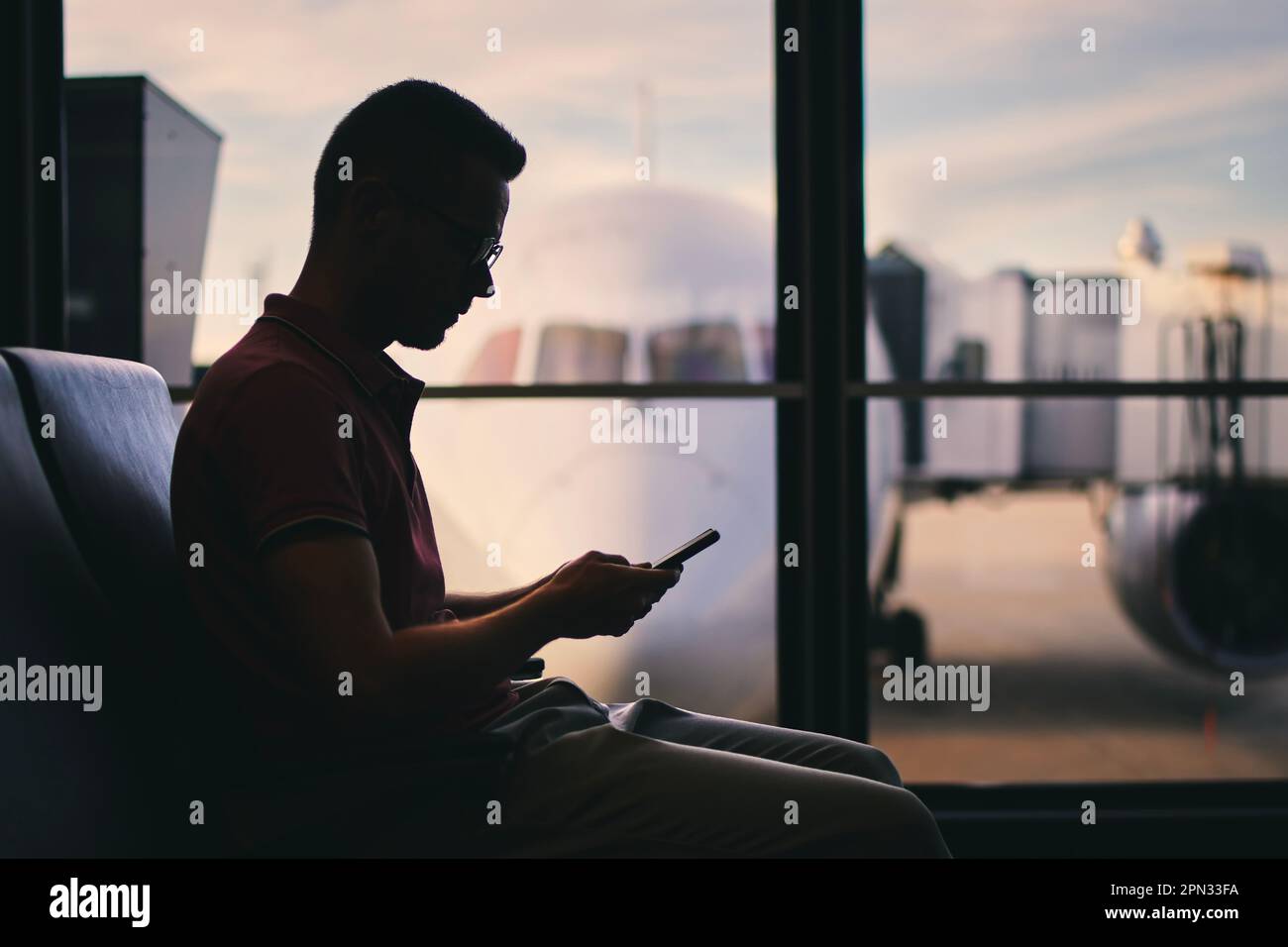Il viaggiatore utilizza il telefono mentre attende il volo. Silhouette di uomo in attesa dalla finestra del terminal dell'aeroporto dietro il quale è l'aereo. Foto Stock