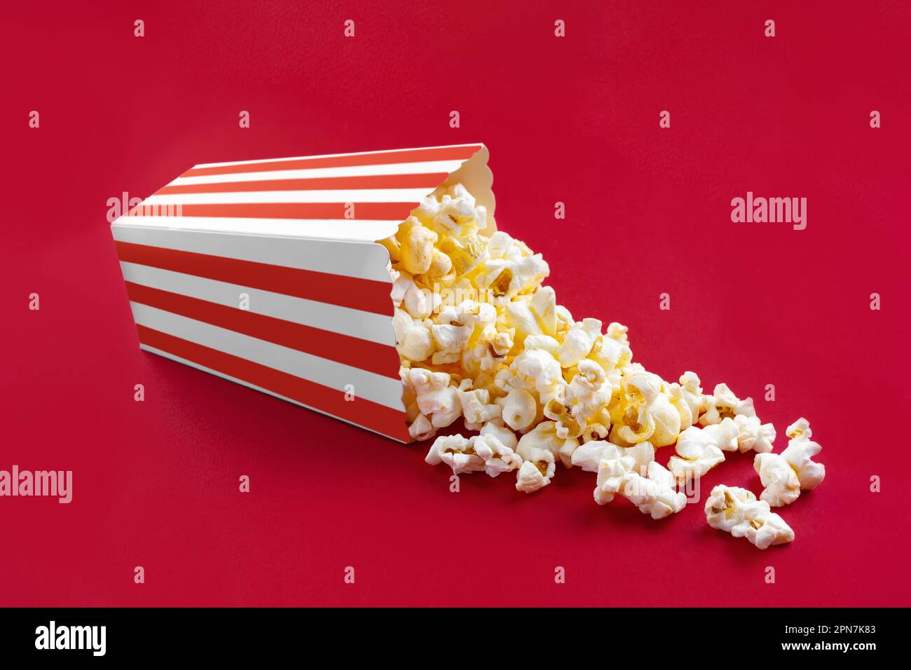 Gustoso popcorn al formaggio che cade da un secchio di cartone a strisce rosse, isolato su sfondo rosso. Dispersione di grani di popcorn. Film, cinema e intrattenimento Foto Stock