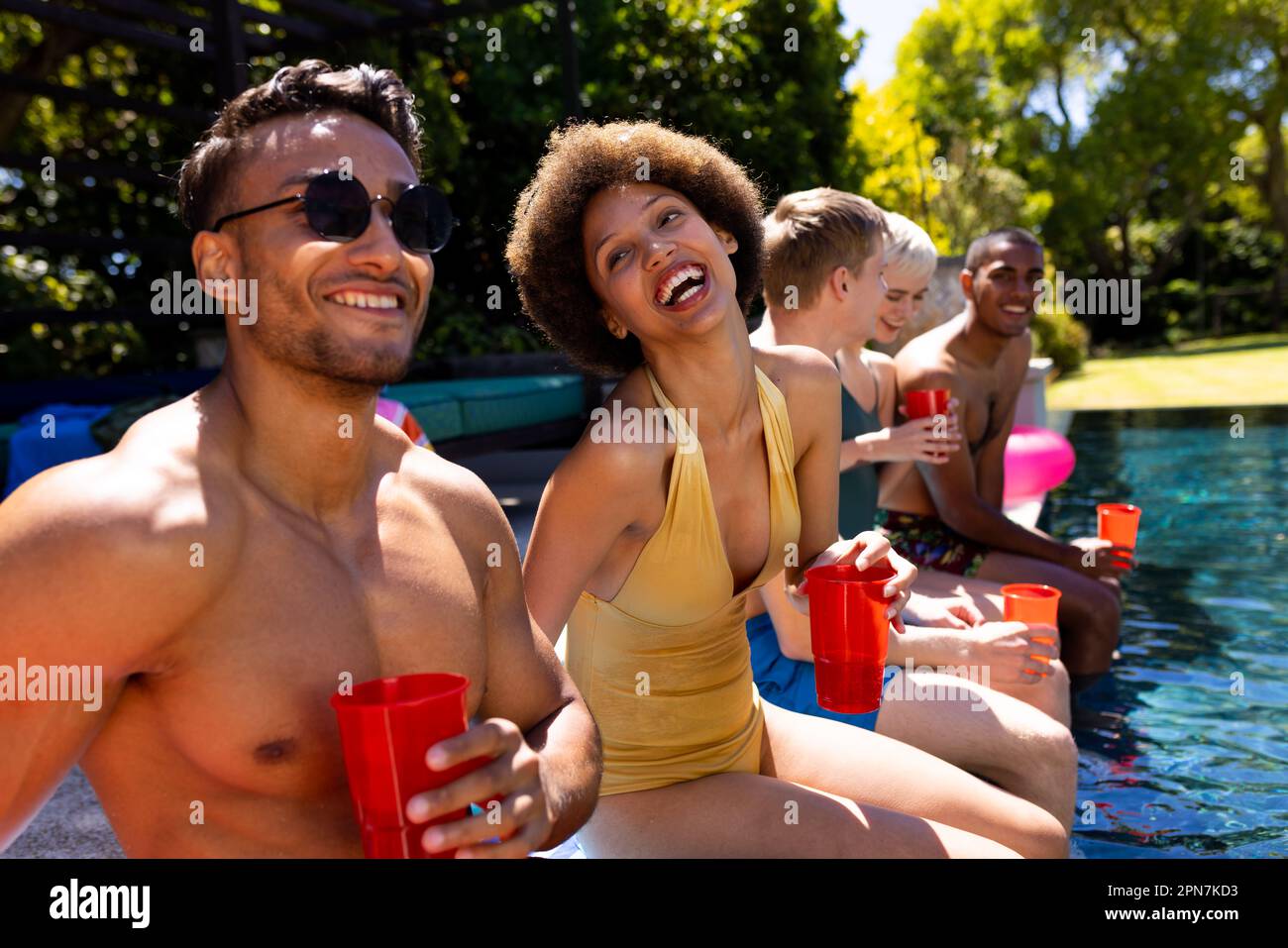 Felice gruppo diversificato di amici che hanno partito in piscina, tenendo tazze di plastica in giardino Foto Stock