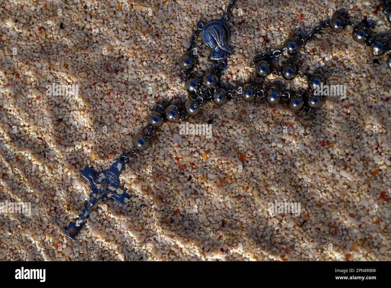 Un crocifisso d'argento e un rosario con perle parzialmente sepolte nella sabbia. Concetto di religione, fede e spiritualità. Foto Stock