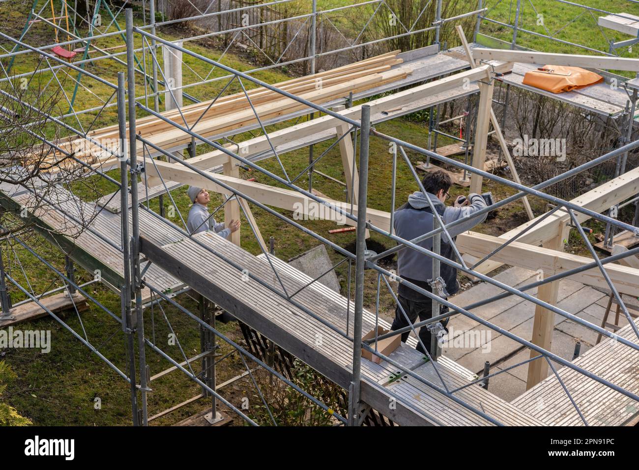 Due giovani falegnami lavorano in cantiere. Un falegname esegue un foro in una trave con un trapano elettrico, e si trova su una ringhiera. Seconda carpina Foto Stock