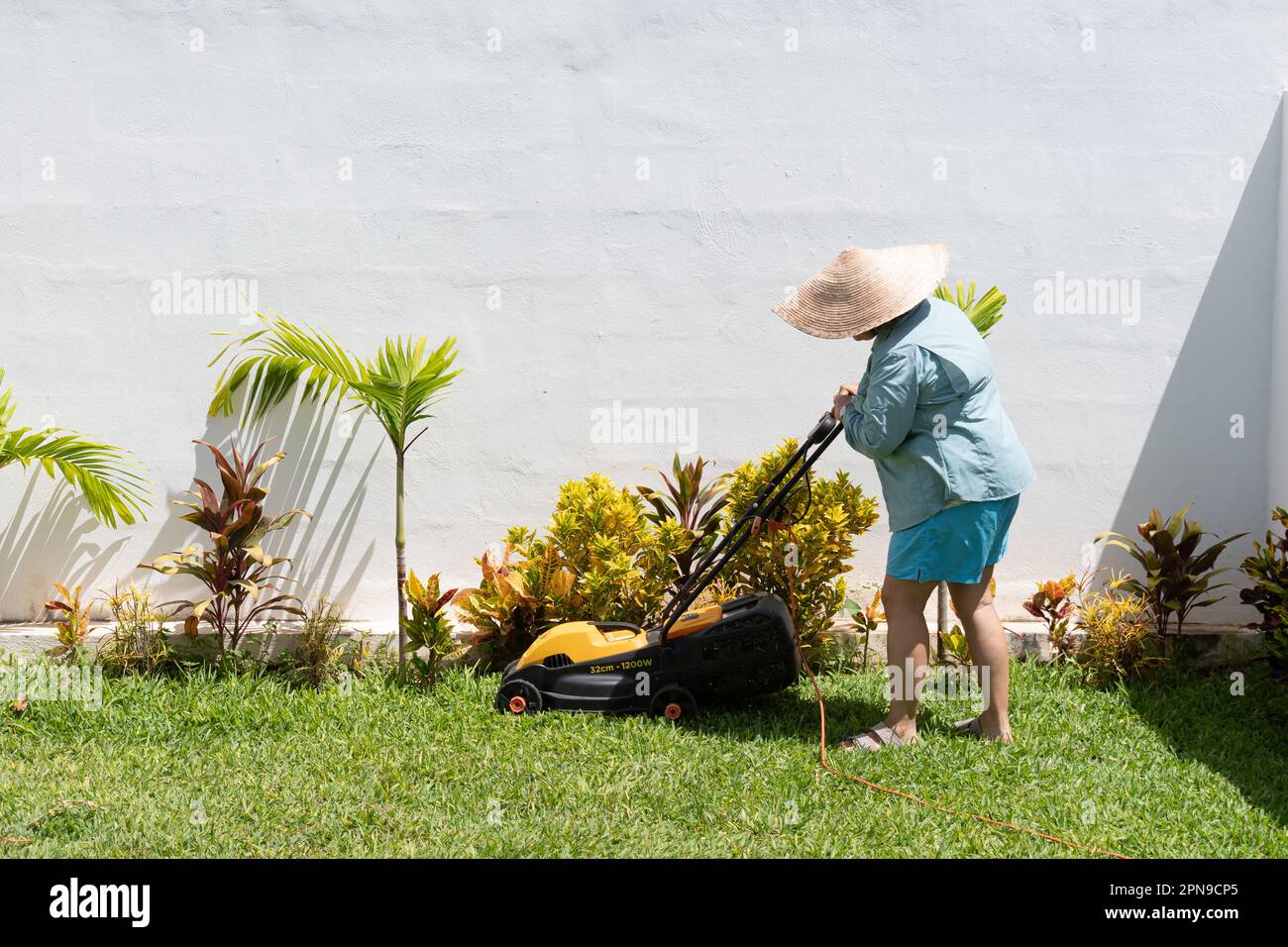 Donna che indossa pantaloncini blu, camicia a maniche lunghe e cappello, taglia l'erba nel giardino della sua casa con un rasaerba. Foto Stock