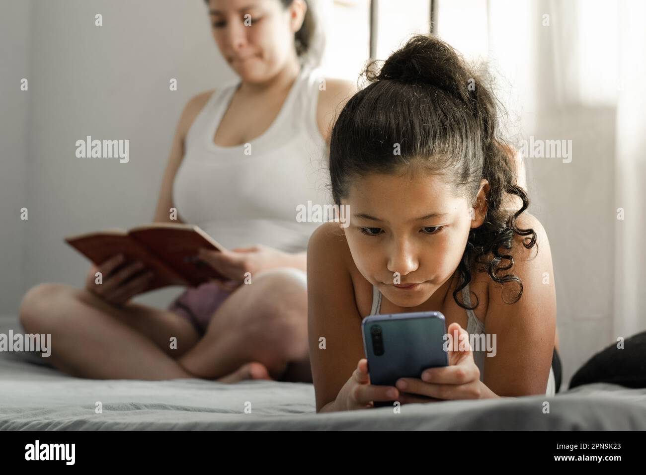 piccola bruna, sdraiata sulla schiena guardando il suo telefono cellulare mentre sua madre legge un libro sullo sfondo Foto Stock