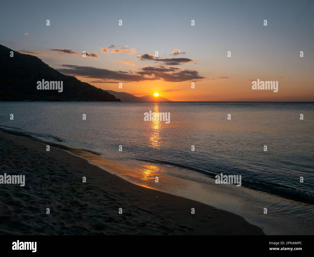 Il sole tramonta dietro colline silhouette lungo la costa di un'isola tropicale, la sua luce si riflette sulla superficie di un mare calmo. Filippine. Foto Stock