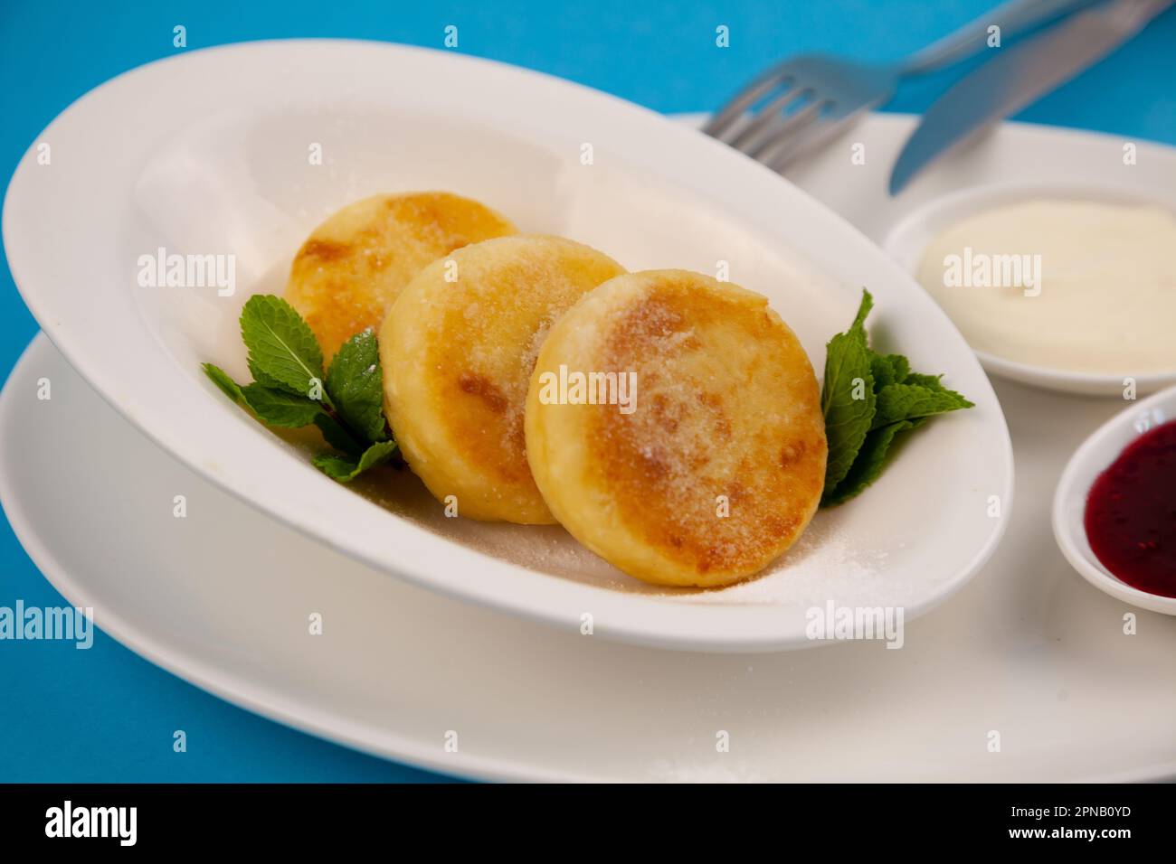 Frittelle di formaggio ricotta rotonda in un piatto bianco su sfondo blu. Cibo prima colazione. Cibo sano. Salsa di panna acida e menta. Primo piano vista dall'alto. Pasto dessert. Frittelle di ricotta al formaggio ricotta. Foto Stock