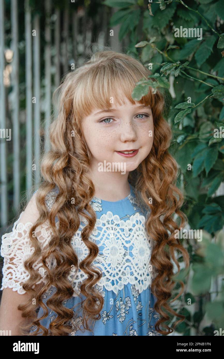 Ritratto di una ragazza con lunghi capelli biondi per strada. Una ragazza di 9-11 anni su uno sfondo di piante verdi, sorridendo e guardando la macchina fotografica. Foto Stock