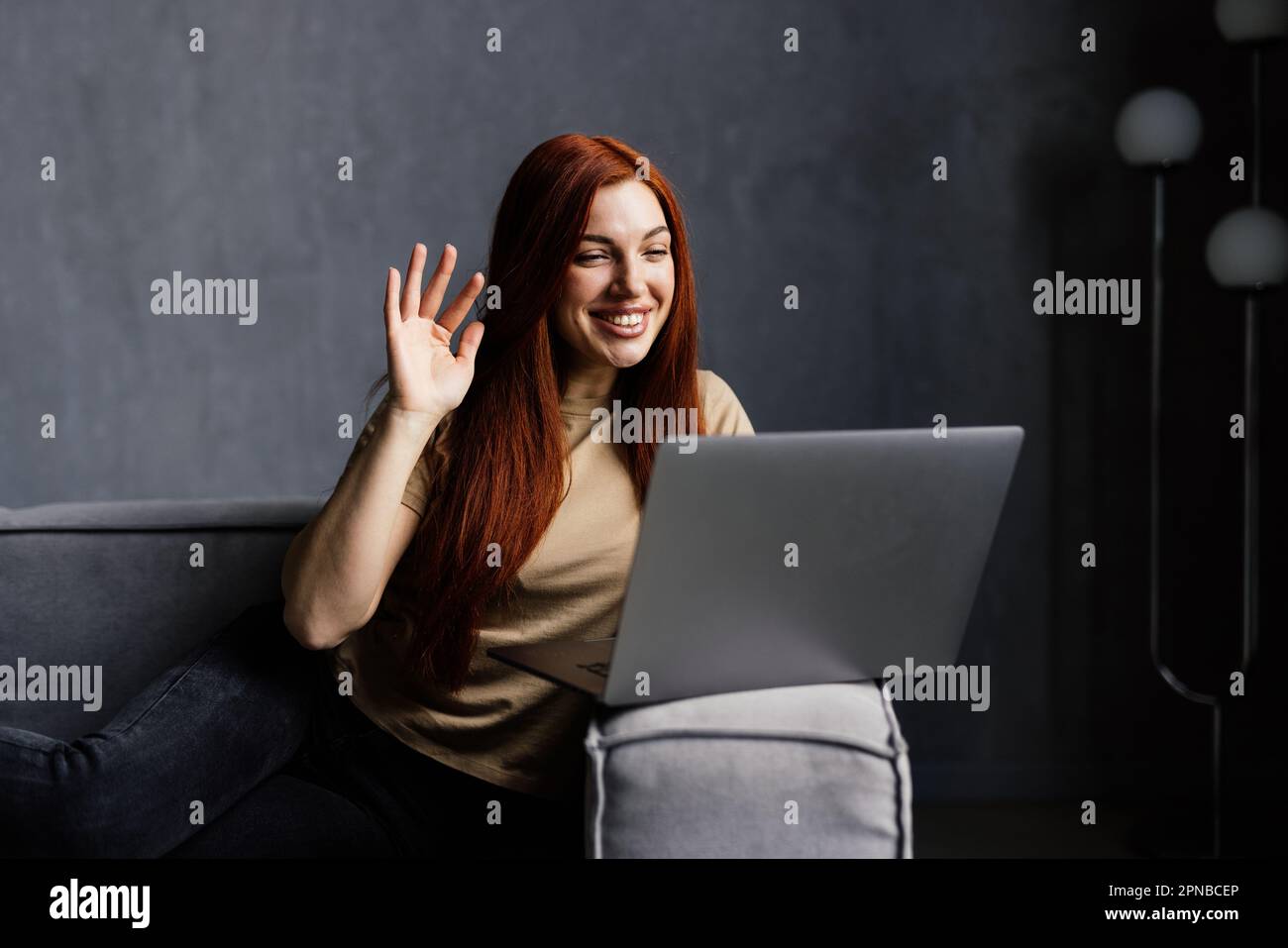 Felice ragazza videochiamata via computer portatile, salutando la webcam, seduto sul divano nel soggiorno. Donna allegra comunicare online, utilizzando moder Foto Stock
