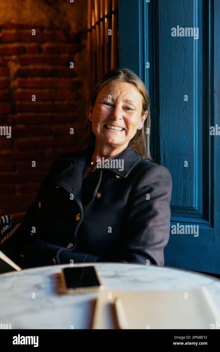 Donna di mezza età positiva in giacca nera guardando la fotocamera mentre si siede al tavolo con smartphone vicino alla porta blu Foto Stock