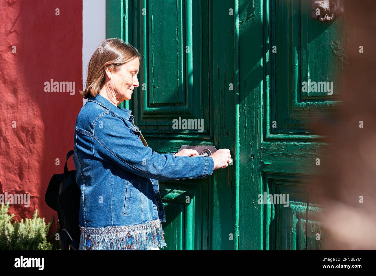 Vista laterale di una donna matura in abiti casual con zaino che lascia la casa e blocca la porta verde di legno del vecchio edificio sulla strada Foto Stock