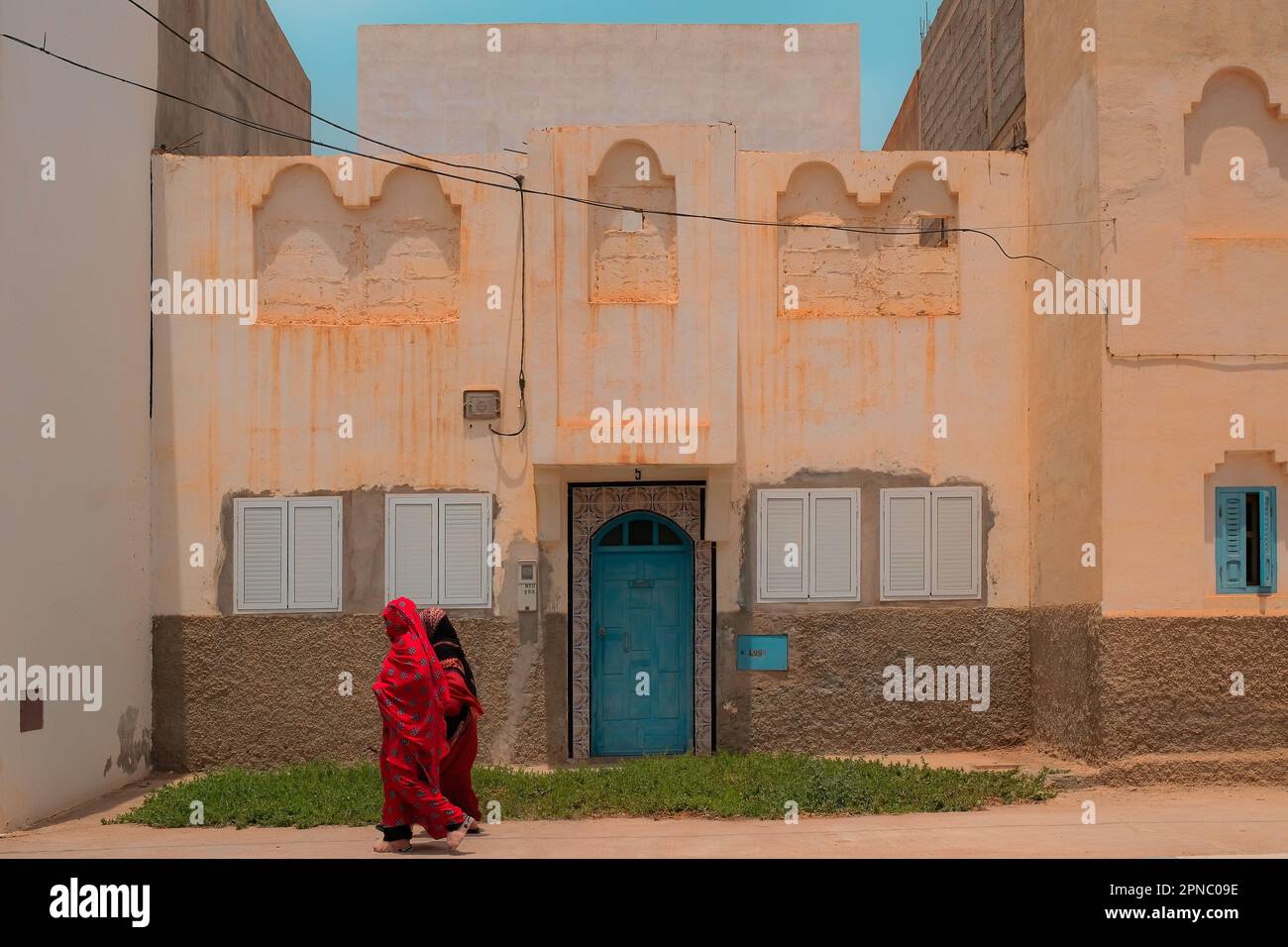 Due donne musulmane locali ricoperte di Abaya rossa camminano davanti ad una vecchia casa su un marciapiede a Sidi Ifni, Marocco durante il Ramadan. Abbigliamento religioso tradizionale. Foto Stock