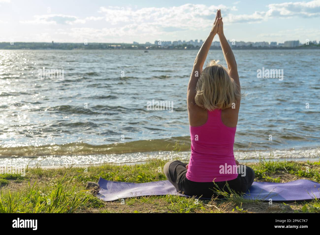 Una donna anziana seduta nella posizione del loto e sollevando le mani in su e guardando il lago, una vista dal retro. Meditazione. Sport in età avanzata. Foto Stock