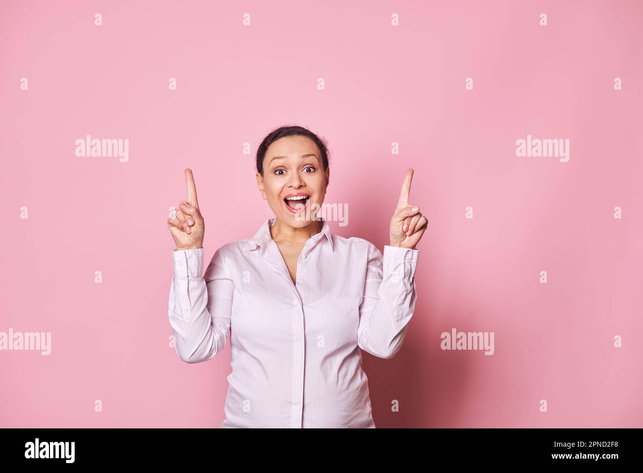 Donna incinta stupita che punta le dita verso l'alto, in posa con la bocca aperta, esprimendo emozioni positive su sfondo rosa Foto Stock
