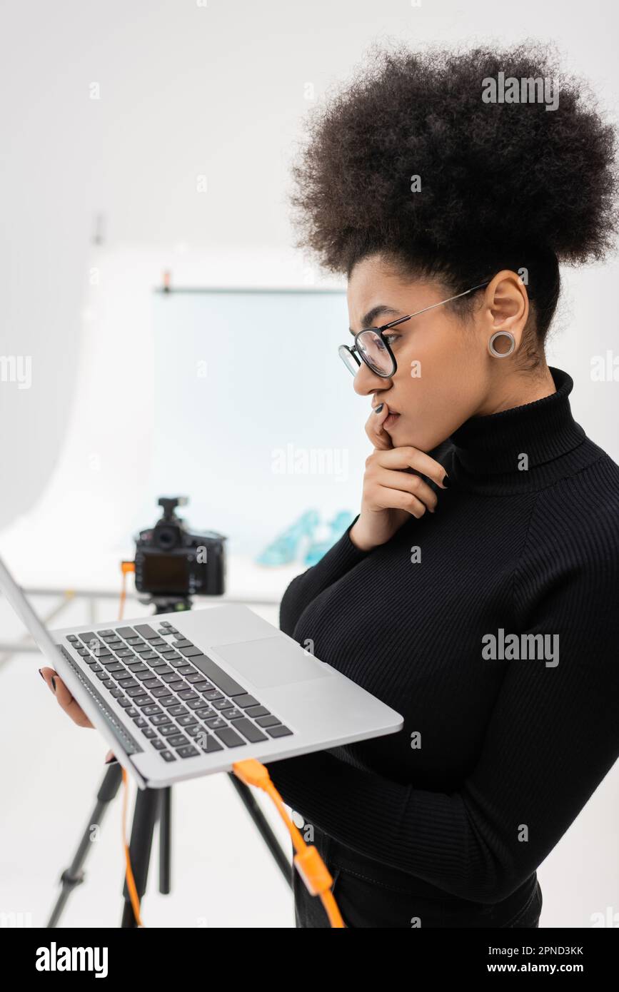 vista laterale di un pensieroso creatore di contenuti afro-americani guardando il portatile in uno studio fotografico sfocato, immagine di scorta Foto Stock