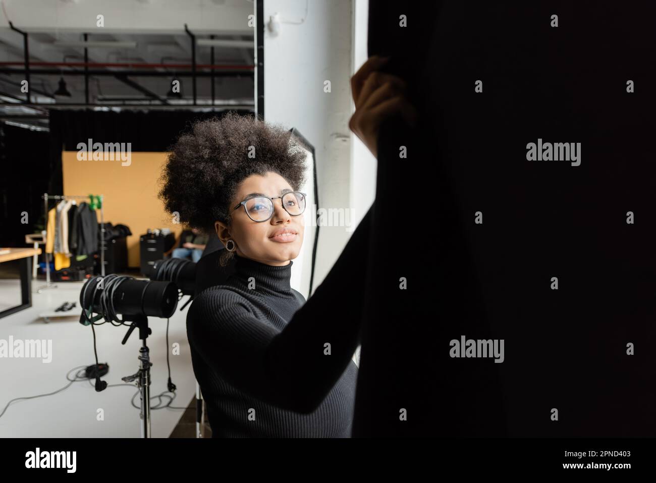 african american content maker in occhiali sorridenti vicino alla tenda nera in studio fotografico, immagine stock Foto Stock