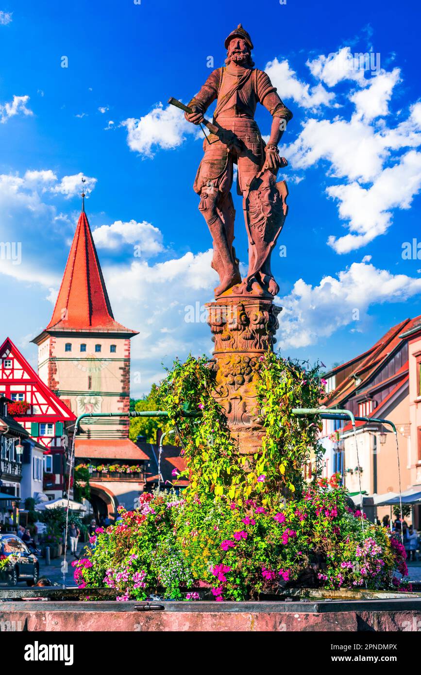 Gengenbach, Germania. Rohrbrunnen fontana, costruita nel 1587, circondata da affascinanti edifici a graticcio e coloratissime composizioni floreali. Forche nere Foto Stock
