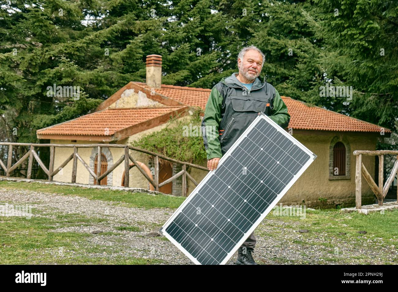 Uomo bearded di mezza età che tiene il pannello solare vicino alla casa nella zona rurale. Energia alternativa, fonti di energia rinnovabile e risorse energetiche sostenibili con Foto Stock