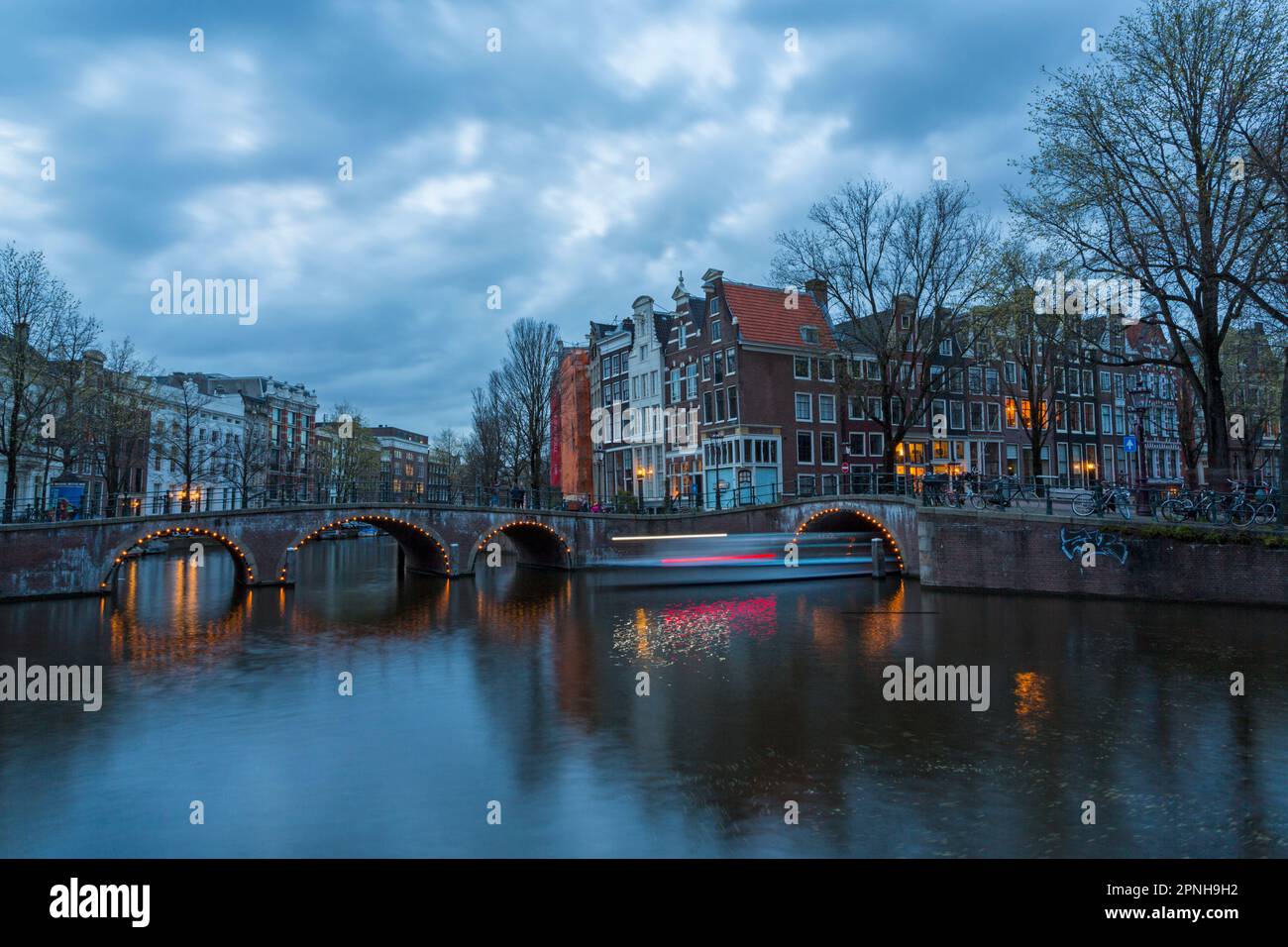 Ponti illuminati al crepuscolo ora blu ad Amsterdam, Olanda, Paesi Bassi nel mese di aprile - lunga esposizione con luci di barca che passano sotto il ponte Foto Stock