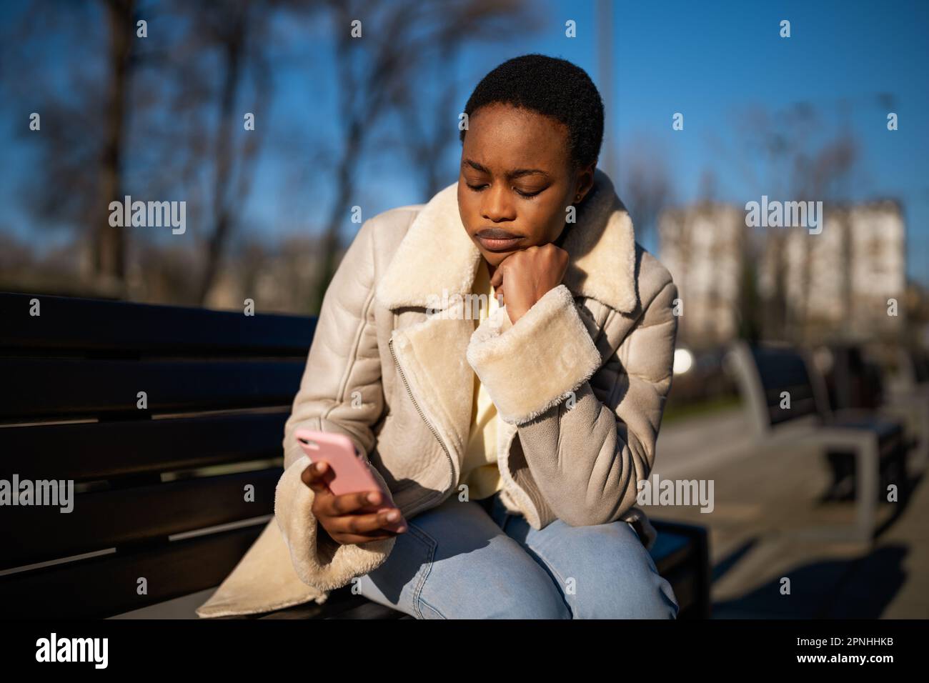 Ritratto all'aperto di una donna nera preoccupata nelle giornate di sole. È seduta su una panchina per strada e la messaggistica al telefono. Foto Stock