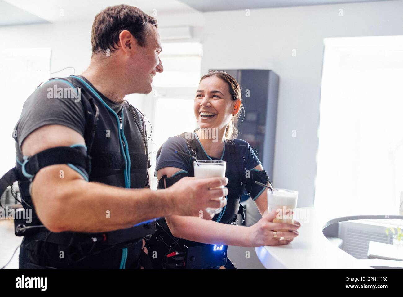 Giovane uomo sorridente e donna in abiti ems comunicare e bere proteine shakes nel bar dello sport. Resto dall'addestramento con gli stimolatori elettrici del muscolo. MO Foto Stock